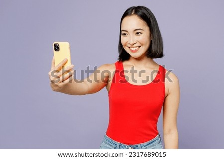 Young woman of Asian ethnicity she wear casual clothes red tank shirt doing selfie shot on mobile cell phone post photo on social network isolated on plain pastel purple background. Lifestyle concept