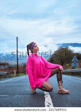 A girl in a pink sweater and a sports headband sitting in the middle of the road on the dividing line, under cloudy weather Royalty-Free Stock Photo #2317689511