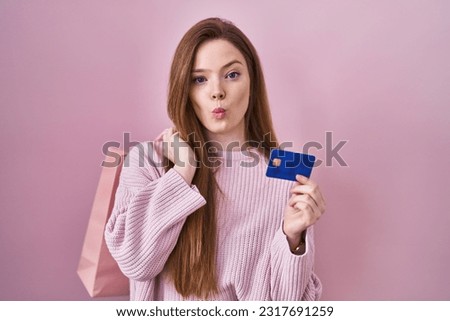 Young caucasian woman holding shopping bag and credit card making fish face with mouth and squinting eyes, crazy and comical. 