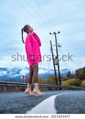 A girl in a pink sweater and a sports headband standing in the middle of a mountain road on the median strip, under cloudy weather Royalty-Free Stock Photo #2317691477