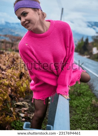A girl in a pink sweater and a sports headband with a long braid climbing over a security barrier on the side of the road, amidst cloudy weather and low clouds in the mountains Royalty-Free Stock Photo #2317694461