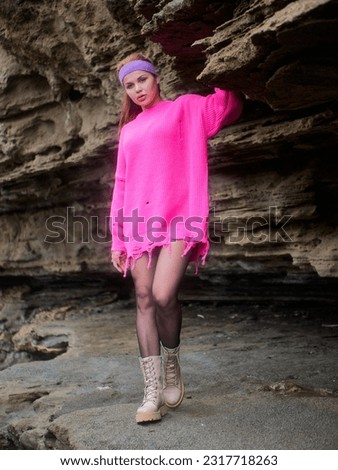 Girl posing in a pink sweater and a sports headband against sharp stone ledges of a cliff, showcasing tectonic layers of time, on a cloudy day
