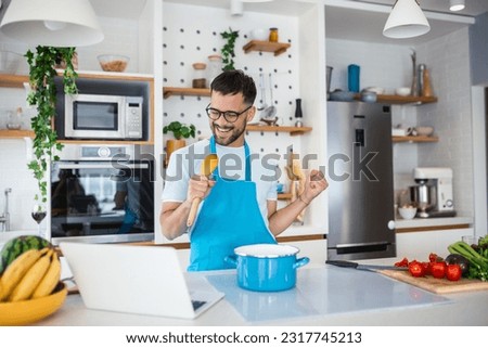 Attractive young man dancing and singing in the kitchen stock photo. happy male holding kitchenware as microphone, listening to music, dancing, doing housework at home, preparing breakfast