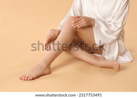 Woman applying body cream onto her smooth legs on beige background, closeup Royalty-Free Stock Photo #2317753495
