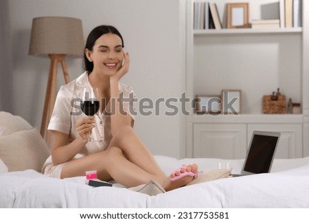 Beautiful young woman with glass of wine giving herself pedicure on bed at home