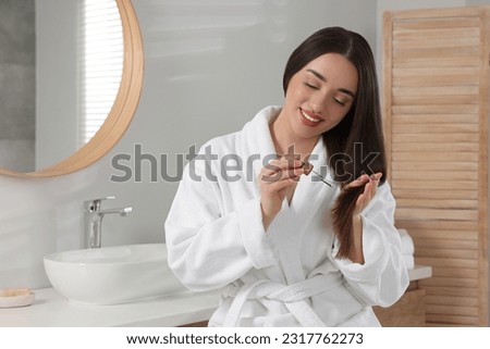 Happy young woman applying essential oil onto hair in bathroom