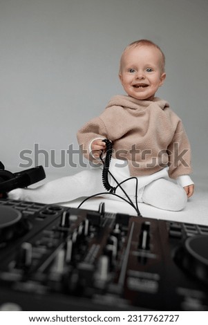 A cute smiling little child in a beige hoodie is sitting on the floor with dj headphones and a dj mixing board. Music and fun. Isolated on grey background.