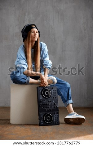 A young pretty long haired DJ girl in a blue sweater, jeans and a black baseball cap is posing while sitting on a white cube. A black DJ mixing console stands nearby. Studio shot, gray background.