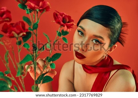 spring style photography, charming asian woman with brunette hair and bold makeup touching green leaves on roses and looking at camera on red background, generation z