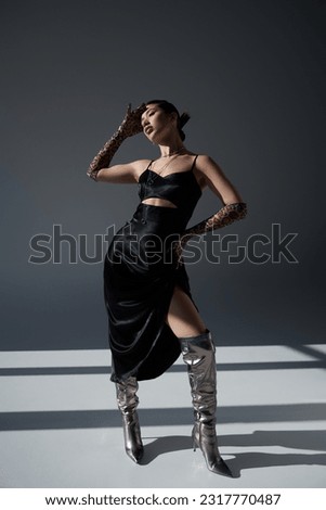 full length of alluring asian woman in silver boots, black fashionable dress and animal print gloves standing with hand on hip on dark grey background with lighting, trendy fashion concept