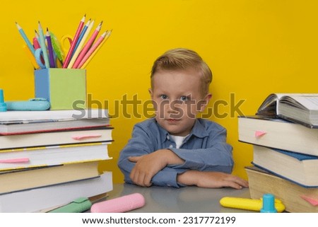 A boy sits at a school desk among books and school supplies on a yellow background copy space