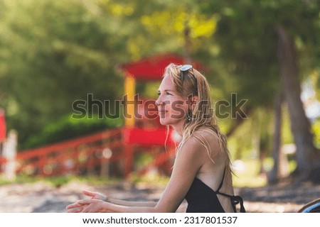Happy woman on tropical beach sitting in swimsuit close shot looking off, vacationing with red lifeguard hut in background on sunny day in shade, relaxing 