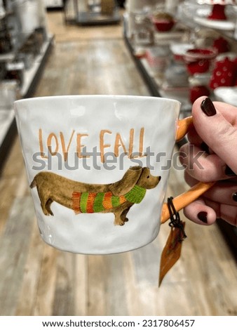 A woman holding a coffee mug with a dachshund on it. The dog is wearing a scarf and the cup says "Love Fall".