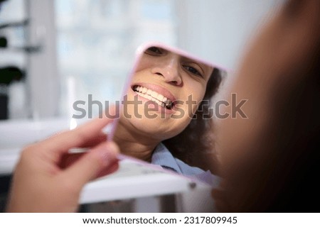 Close-up reflection in the cosmetic mirror of a pretty woman, female patient sitting in dental chair, admiring her beautiful smile and teeth after teeth bleaching procedure in dentistry clinic. Royalty-Free Stock Photo #2317809945