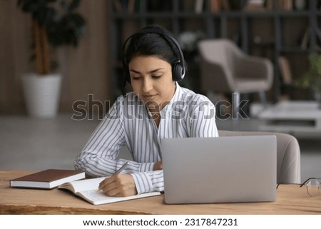 Young indian woman take part in studying online use headphones pc handwriting learning material from virtual education event to copybook. Ethnic female student take notes from online training webinar Royalty-Free Stock Photo #2317847231