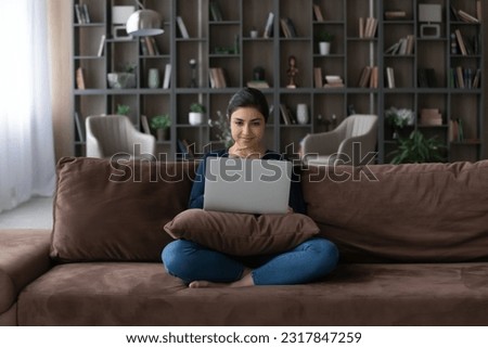 Young indian woman sit cross legged on couch enjoy online date create answer on message from boyfriend at social network. Curious hindu female read interesting information at blog website on pc screen Royalty-Free Stock Photo #2317847259