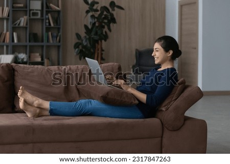 Smart job. Side view of cheerful barefoot lady spend time on weekend with laptop rest on cozy couch at modern living room. Happy indian teenage female enjoy freelance work on pc from home
