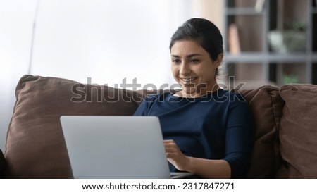Happy young indian lady enjoy weekend rest on sofa chat by video call using wifi internet on laptop watch good comedy movie. Joyful teen female relax at home read funny blog article on computer screen