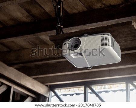 A white projector hangs on the brown wood ceiling of an old wooden house building. Video presentation, slide projector machine technology for business multimedia in local school.