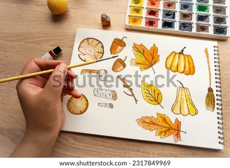 Closeup of women's hand with brush drawing on notepad. Creation process of watercolor painting. Autumn leaves and pumpkins.