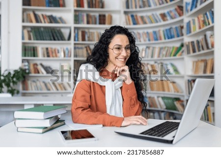 Female student with laptop reading online video course, Latin American woman smiling and satisfied with independent online learning sitting inside university campus in library.