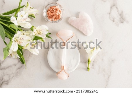 A professional rose quartz massager for facial care at home lies on a white marble round podium next to gua sha and sea salt in a bowl. Top view