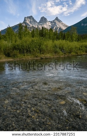 Portrait view of Three Sisters in Canmore seen at golden hour, sunset on blue sky day, afternoon with calm, peaceful reflection in water below famous, tourist, tourism mountains, area summer.   Royalty-Free Stock Photo #2317887251