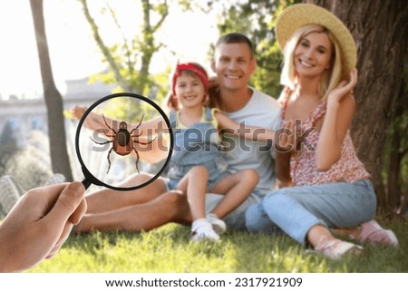 Family sitting near tree outdoors and don't even suspect about hidden danger in green grass. Woman showing tick with magnifying glass, selective focus Royalty-Free Stock Photo #2317921909
