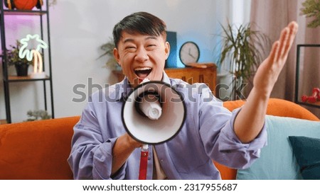 Asian handsome man talking with megaphone, proclaiming news, loudly announcing advertisement, warning using loudspeaker to shout speech, real estate sale at home. Chinese guy sitting on sofa in room Royalty-Free Stock Photo #2317955687