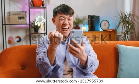 Happy asian man use mobile smartphone typing browsing say Wow yes found out great big win good news celebrate lottery jackpot doing winner gesture. Chinese adult guy at home in room sitting on sofa Royalty-Free Stock Photo #2317955739