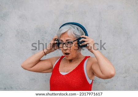 Amazed elderly woman touching headphones against concrete wall