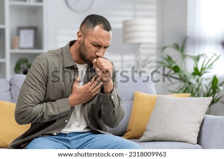 Young African American man sitting on the couch at home and coughing. Suffers from an attack of asthma, allergies. He holds his chest, covers his mouth with his hand. Royalty-Free Stock Photo #2318009963