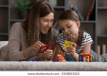 Cute girl and happy mom engaged in learning game together, playing construction toys, building blocks, connecting pieces, plastic details, relaxing on clean carpet on heating floor. Family playtime Royalty-Free Stock Photo #2318023323