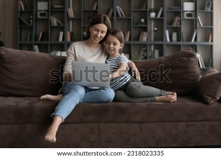 Happy millennial mom and gen Z daughter girl relaxing on couch at home, using laptop, watching movie online, making video call, shopping on internet, enjoying leisure time with digital device.