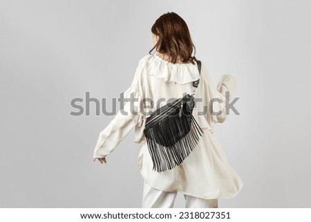 White Woman from Behind Wearing White Clothes and Crossbody Black Leather Belt Bag with Boho Style Decorative Fringe over Grey Background Royalty-Free Stock Photo #2318027351
