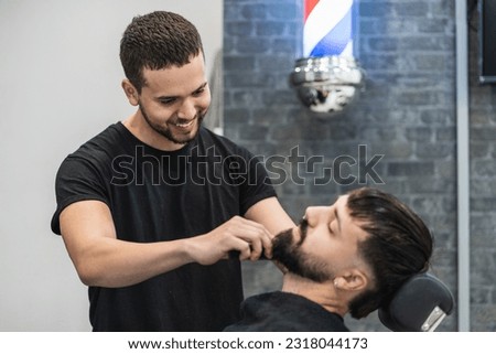 Smiling barber combing and trimming his client's beard.