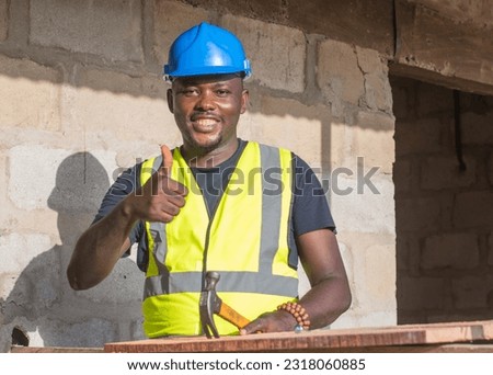 A happy male African construction worker with a blue safety helmet on his head, doing thumbs up gesture, and working with an hammer on a building site in Nigeria Royalty-Free Stock Photo #2318060885