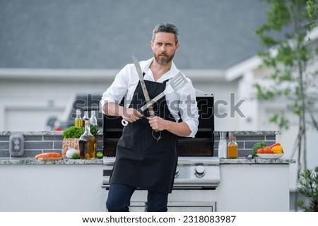Man at a barbecue grill. Male cook preparing barbecue outdoors. Bbq meat, grill for picnic. Roasted beef. Cook preparing barbeque in the house yard. Barbecue and grill. Cook using barbecue tongs. Royalty-Free Stock Photo #2318083987