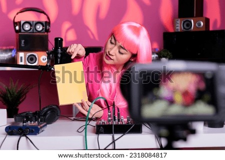 Asian influencer holding notebook advertising product in front of camera while recording school supplies review using broadcast equipment. Content creator with pink hair live streaming