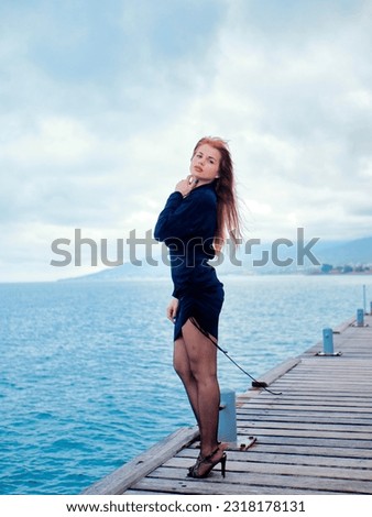 Red-haired girl in a black dress standing on a wooden pier, with the coastline of the sea and mountains in the background Royalty-Free Stock Photo #2318178131