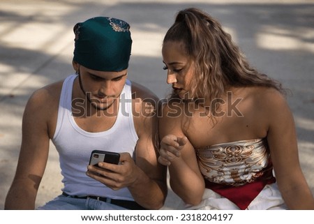 Young, non-conformist Latino and Hispanic boy and girl couple sitting on a bench checking social networks on their cell phone. Mobile concept, smartphone, networks, applications, dependence.