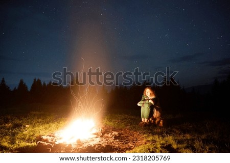 A young female traveler sits by the campfire near her tent, enjoying a moment of relaxation beneath the beautifully starry sky. As the crackling flames provide warmth and comfort