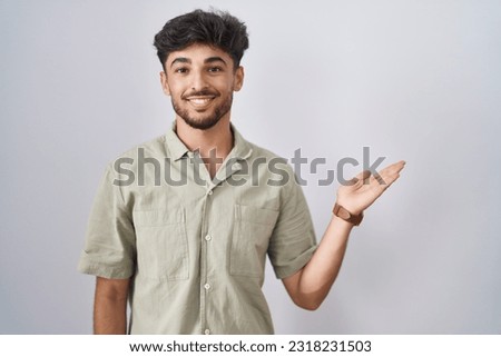 Arab man with beard standing over white background smiling cheerful presenting and pointing with palm of hand looking at the camera. 