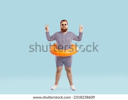 Funny chubby man with inflatable circle for swimming at waist shows V-sign on light blue background. Full length of serious humorous redhead bearded young man in striped leotard who show peace v-sign.