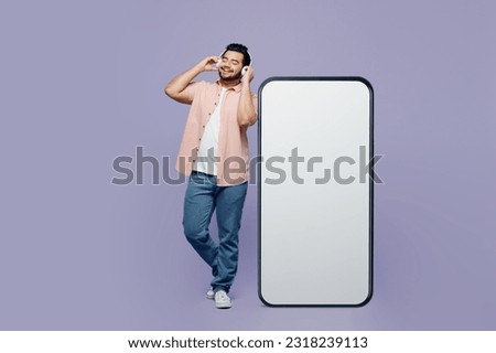 Full body young Indian man wear pink shirt white t-shirt casual clothes big huge blank screen mobile cell phone smartphone with area listen music headphones isolated on plain pastel purple background