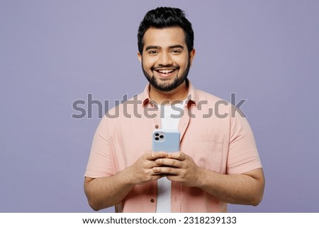 Young smiling happy Indian man he wear pink shirt white t-shirt casual clothes hold in hand use mobile cell phone look camera isolated on plain pastel light purple background studio. Lifestyle concept Royalty-Free Stock Photo #2318239133