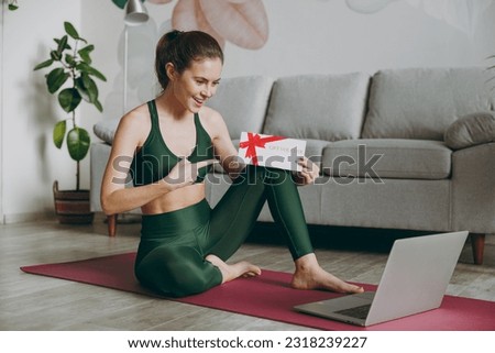 Full body young strong athletic fitness trainer instructor woman wears green tracksuit hold laptop pc computer show gift coupon voucher card train do exercises at home gym indoor Workout sport concept