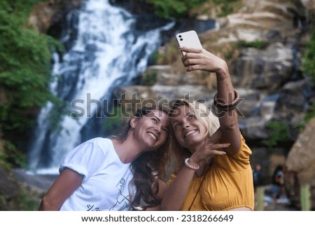 Two happy smiling young women takes selfie at waterfall background. Ladies in casual attire with mobile phone enjoying at tropical nature waterfalls, tropic view. Travel vacation concept. Copy space