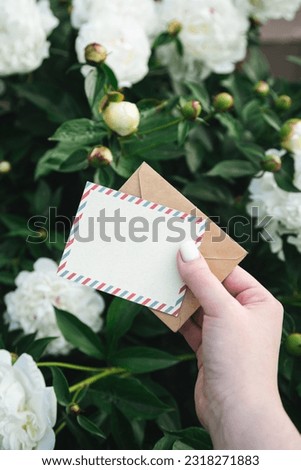 Blank greeting card and a craft envelope in a woman's hand in the garden against the background of a bush of white peonies among the foliage, a festive background with a place for text.