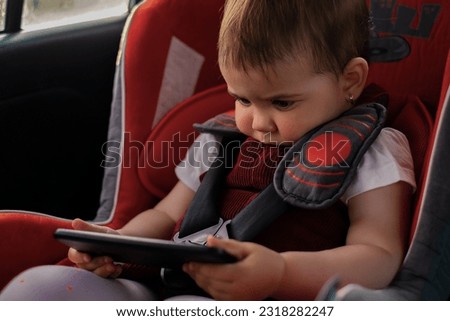 the child is sitting in a car seat, holding a mobile phone in his hands, watching cartoons on the phone, the child is playing on the phone, the entertainment of the child on the journey